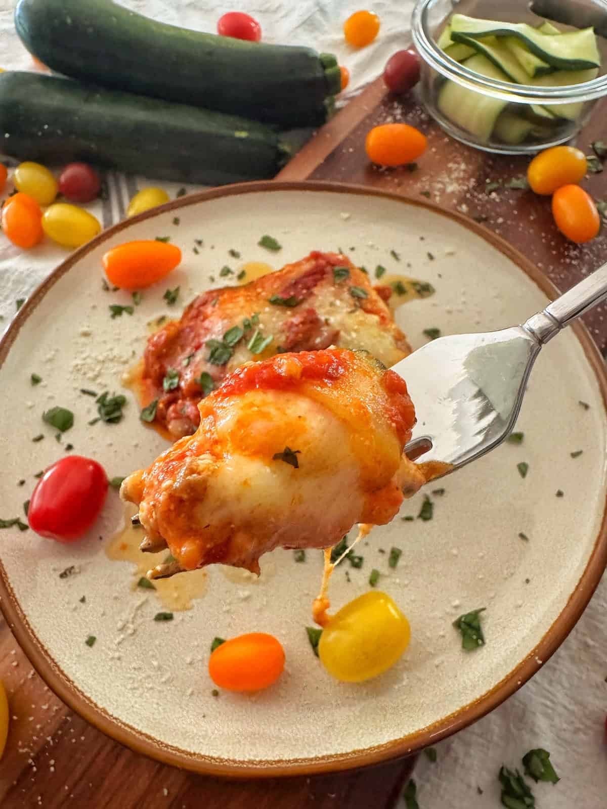 A fork holds a cheesy bite of baked zucchini lasagna over a plate, surrounded by colorful cherry tomatoes, zucchini, and herbs on a wooden board.