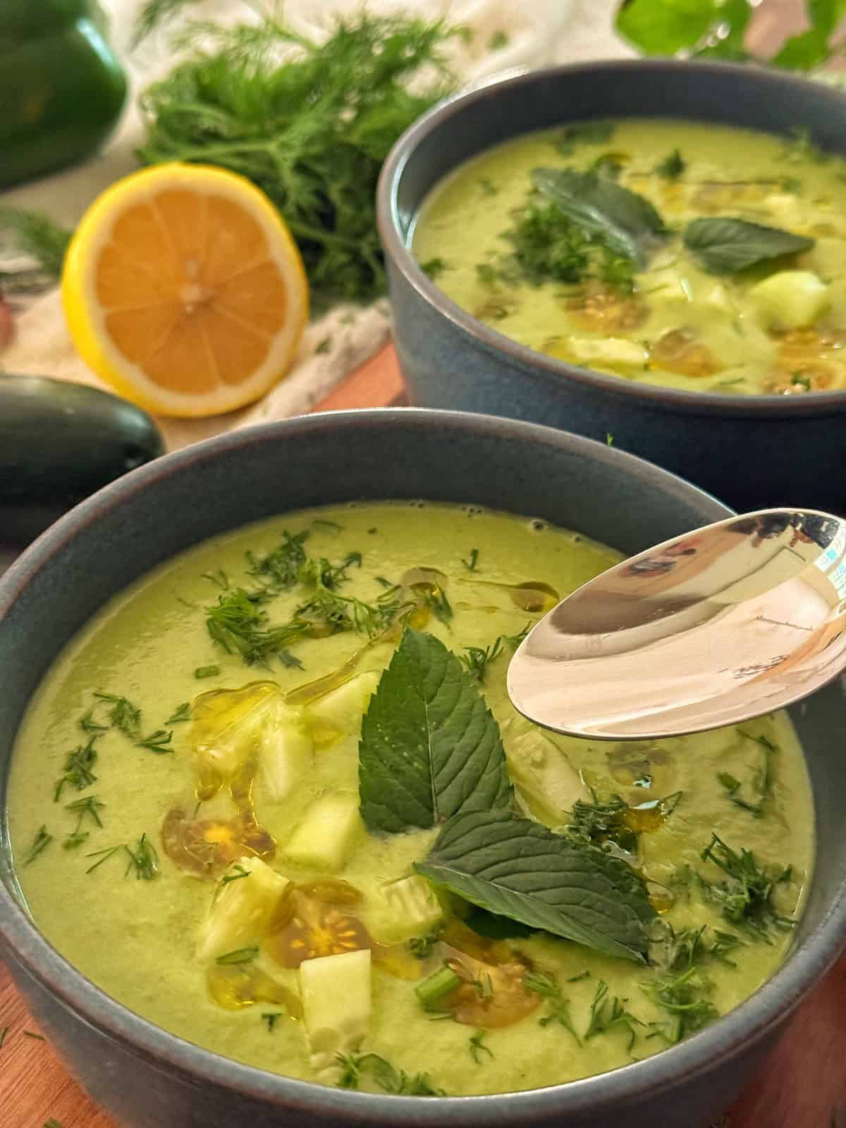 Two bowls of cucumber gazpacho garnished with chopped herbs, cucumber pieces, a mint leaf, and a drizzle of oil. A spoon rests in one bowl. In the background are half a lemon, fresh dill, and a cucumber.