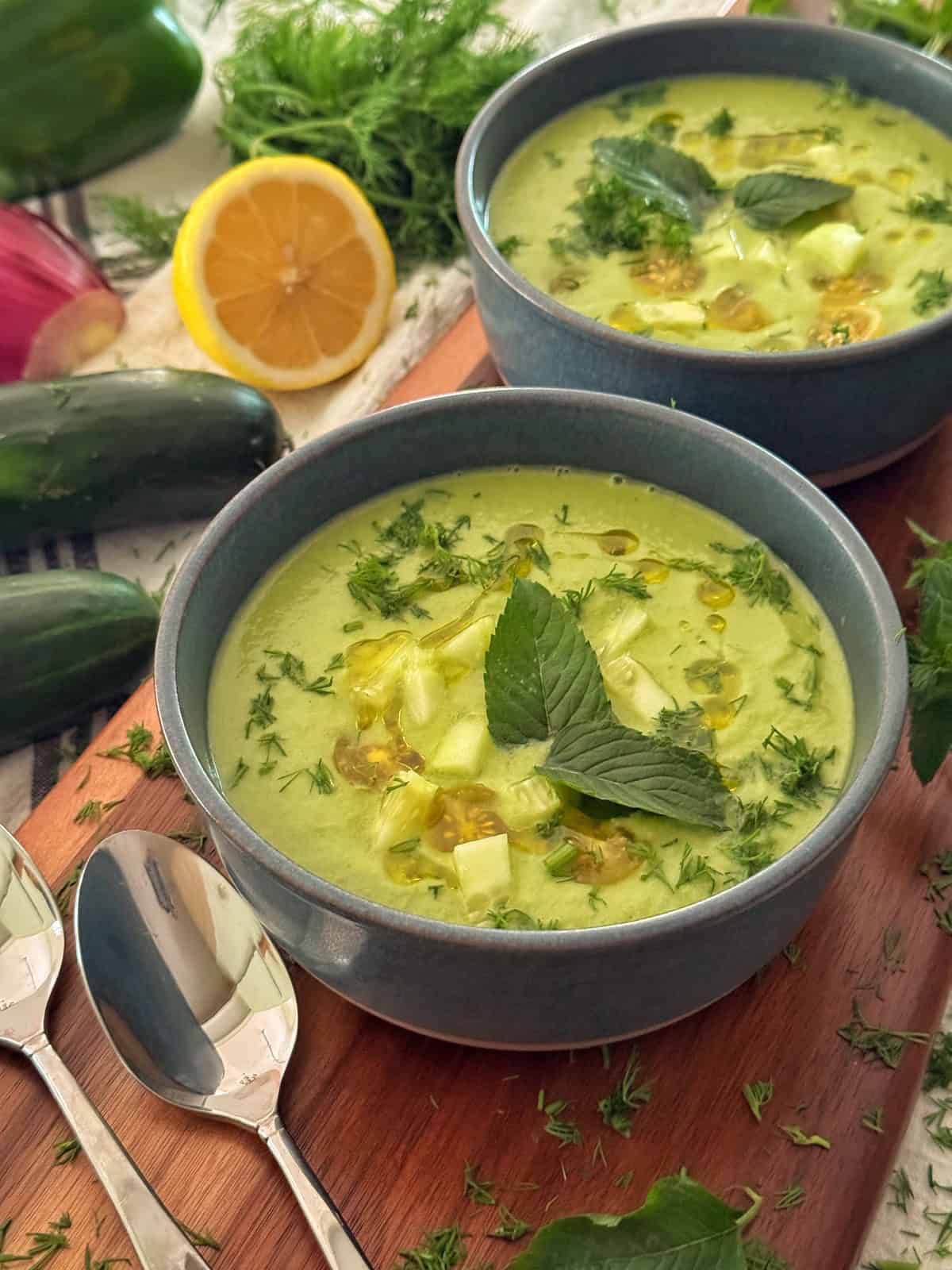 Two bowls of creamy cucumber gazpacho garnished with chopped herbs, diced cucumber, and mint leaves, placed on a wooden board with fresh cucumbers, lemon, dill, and two spoons nearby.