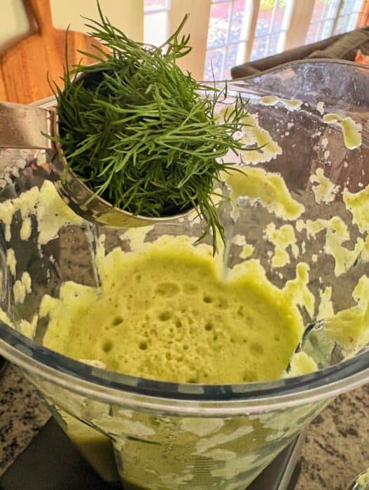 A close-up of a blender containing frothy green liquid to make cucumber soup, with a measuring cup holding fresh dill being added. Sunlight streams through windows in the background.