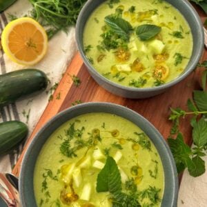 Two bowls of cucumber gazpacho garnished with chopped cucumber, yellow tomatoes, fresh herbs, and a drizzle of olive oil, surrounded by cucumbers, lemon, fresh dill, and mint on a wooden surface.