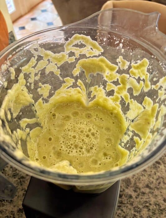 A blender filled with a frothy, light green mixture sits on a kitchen countertop. Some of the mixture is splattered along the sides of the container. The background shows a glimpse of a kitchen area.