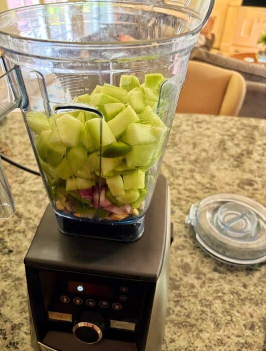 A blender on a granite countertop is filled with chopped green cucumbers, red onions, and other vegetables to make cucumber soup. The blender lid is placed nearby. The background shows part of a kitchen and living room.