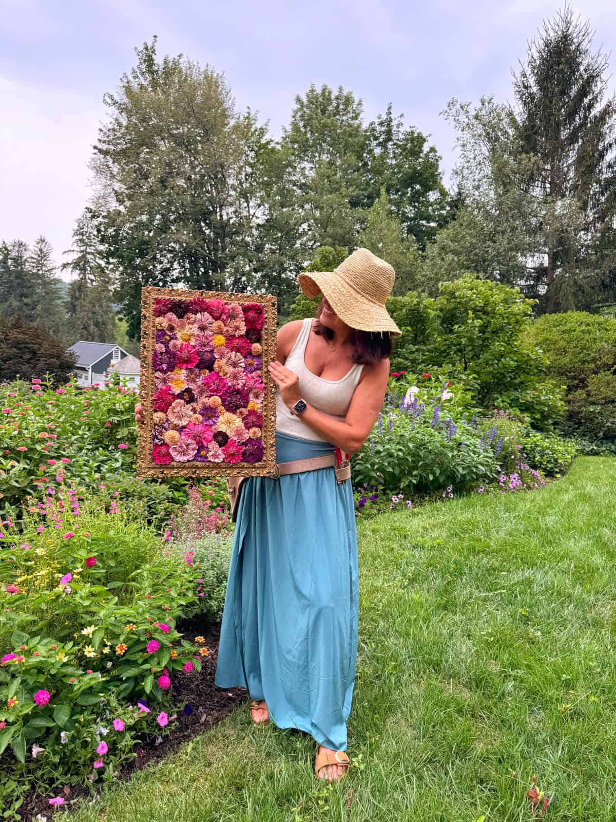 A woman in a wide-brimmed hat and blue skirt stands in a garden holding a colorful floral arrangement framed with pink, red, and yellow flowers. Lush greenery and blooming flowers surround her under a cloudy sky.