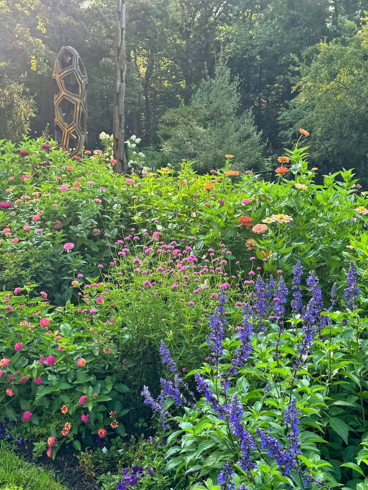 A lush garden with vibrant flowers in pink, orange, and purple blooms, surrounded by green foliage. In the background, a geometric garden sculpture stands among the trees. Sunlight filters through dense greenery.