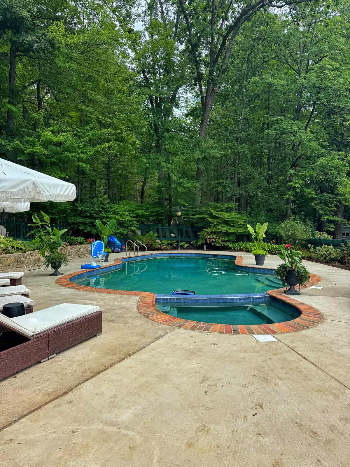 A backyard pool surrounded by brick and concrete decking, lounge chairs, and lush green trees. There are potted plants, a white umbrella, and pool floats by the water, creating a relaxing outdoor space.
