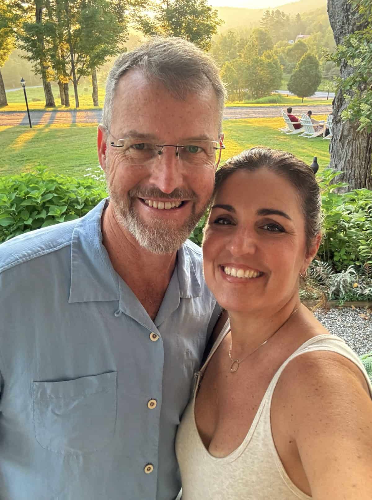 A smiling man wearing glasses and a light blue shirt stands next to a smiling woman in a white tank top. They are outdoors in a sunny, green garden with trees and chairs in the background.