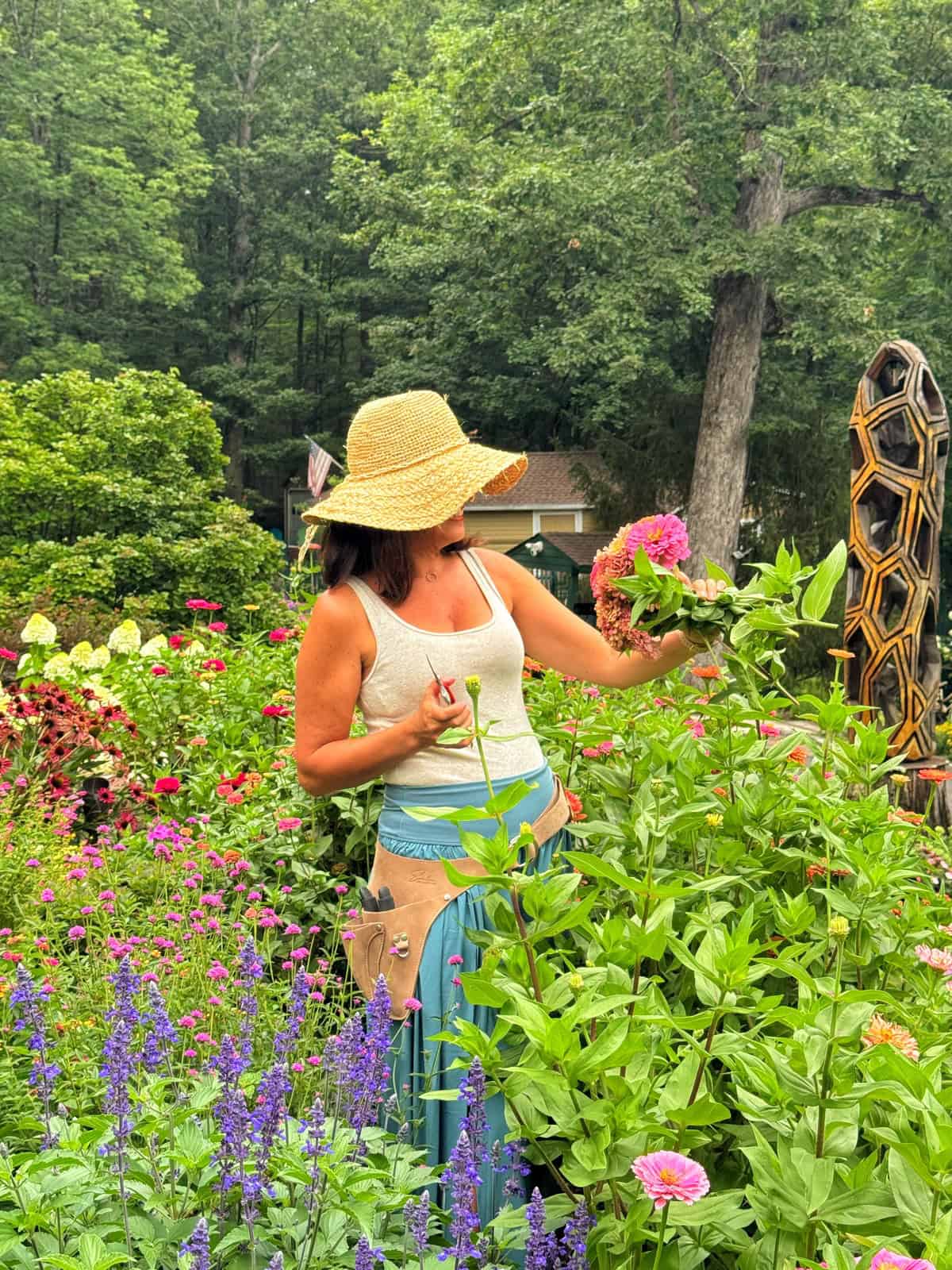 A woman wearing a straw hat and light tank top stands in a colorful garden, holding pink zinnia flowers and pruning scissors, surrounded by lush greenery and blooming flowers.