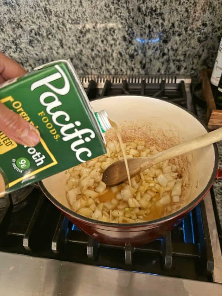 A hand pours Pacific Foods organic vegetable broth into a pot with chopped onions cooking on a stove, while a wooden spoon rests in the pot.