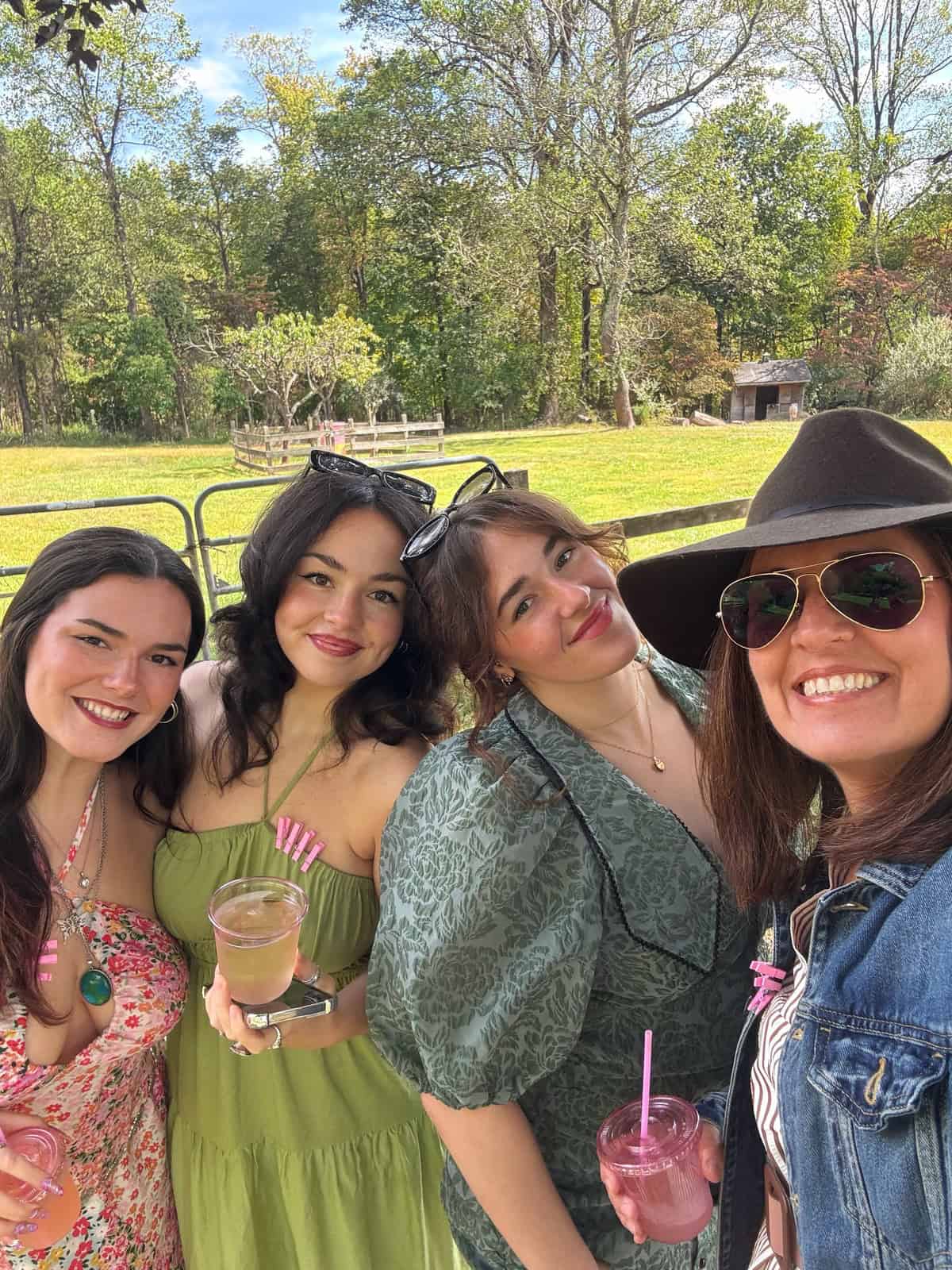 stacy ling and her three daughters smiling outdoors on a sunny day, wearing colorful dresses and holding drinks. Trees and a grassy area are in the background. One woman wears a wide-brimmed hat and sunglasses.
