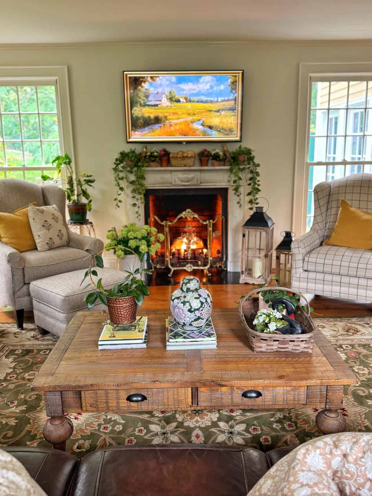 Cozy living room with armchairs, a wooden coffee table holding plants and books, a lit fireplace decorated with greenery, and a countryside painting above the mantel. Large windows let in natural light.