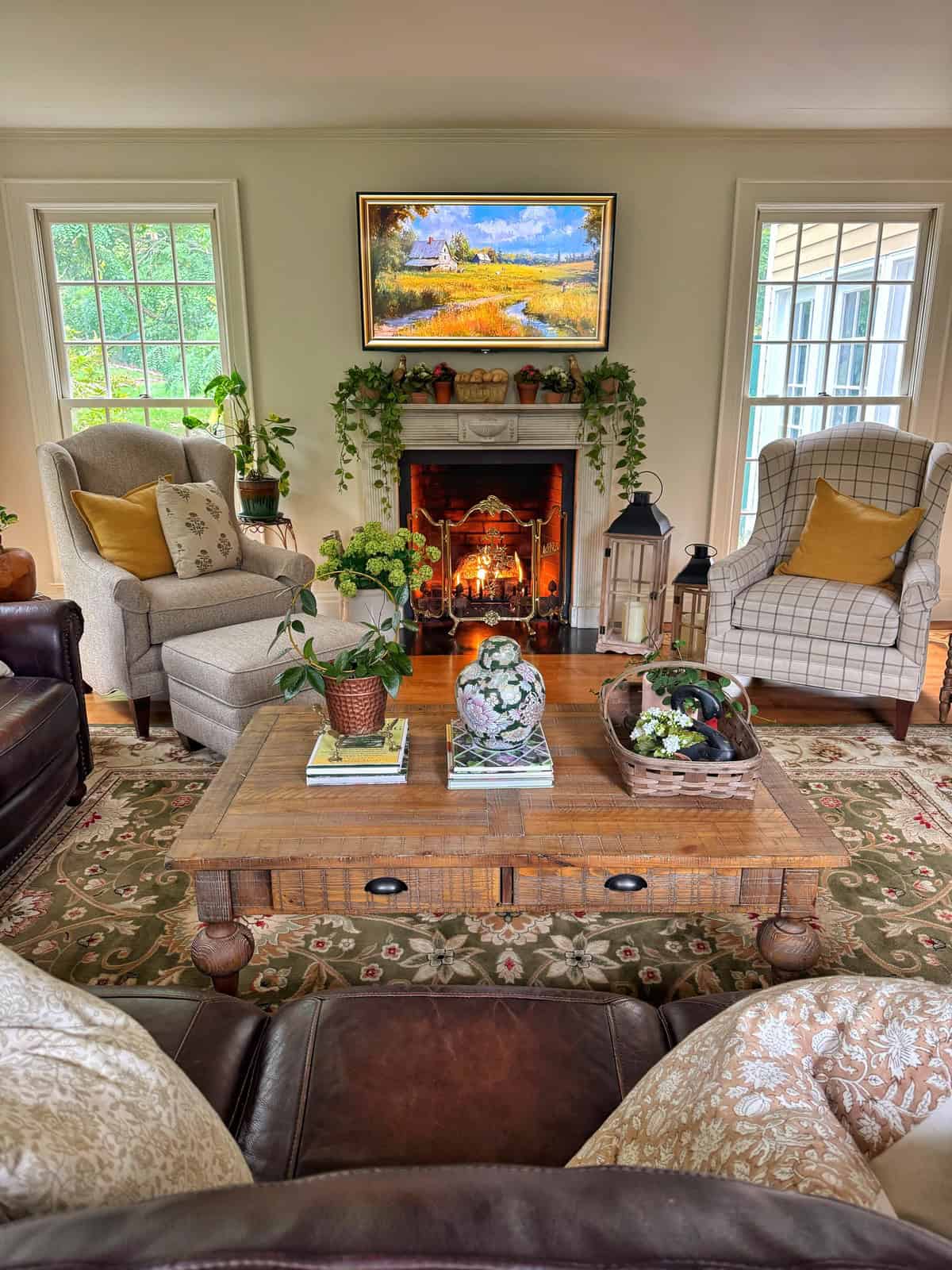 A cozy living room with two armchairs, a fireplace with a fire burning, a wooden coffee table with books and plants, and a large window letting in natural light. A painting hangs above the mantel.