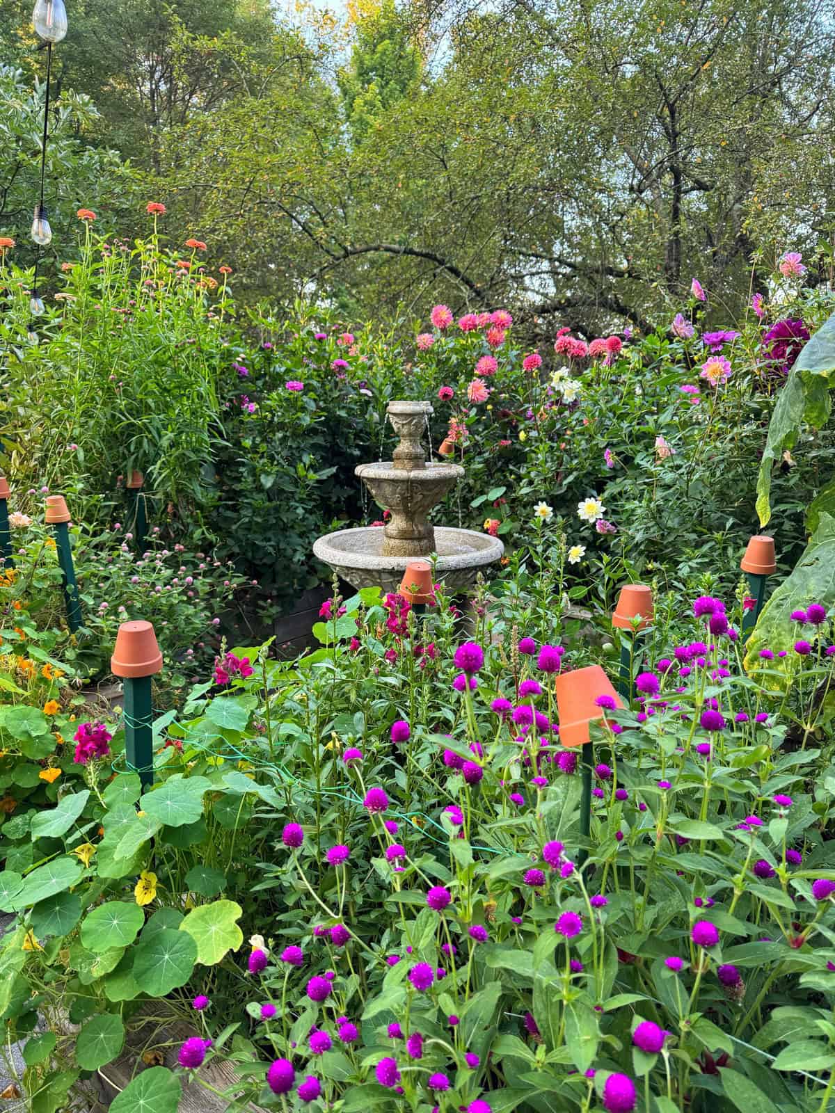 A stone fountain sits in the center of a lush garden filled with blooming flowers in shades of pink, purple, and orange. Green foliage surrounds the fountain, and small pots are placed atop green posts throughout the scene.