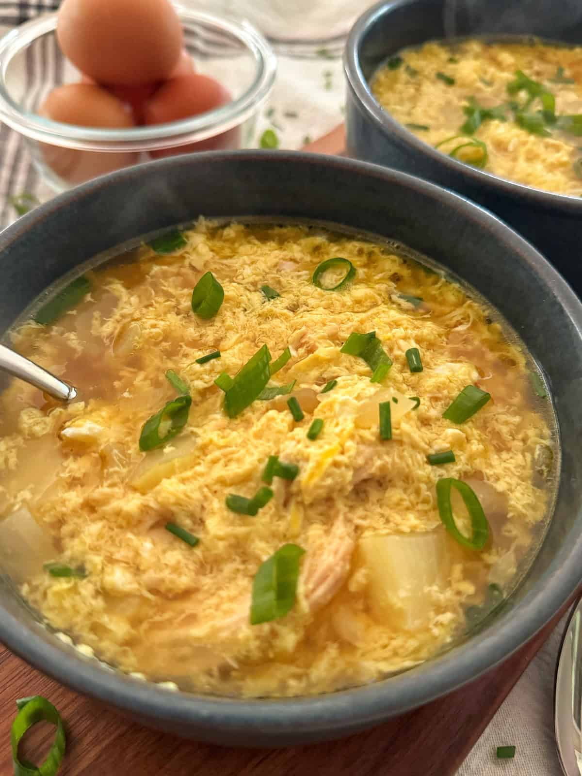 A close-up of a bowl of egg drop soup garnished with chopped green onions, with a spoon in the bowl. Another bowl of soup and a glass bowl with brown eggs are in the background.