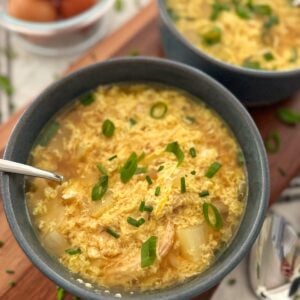 Two bowls of egg drop soup garnished with sliced green onions sit on a wooden surface, with a bowl of eggs and a spoon nearby. The soup contains visible egg ribbons and vegetables.