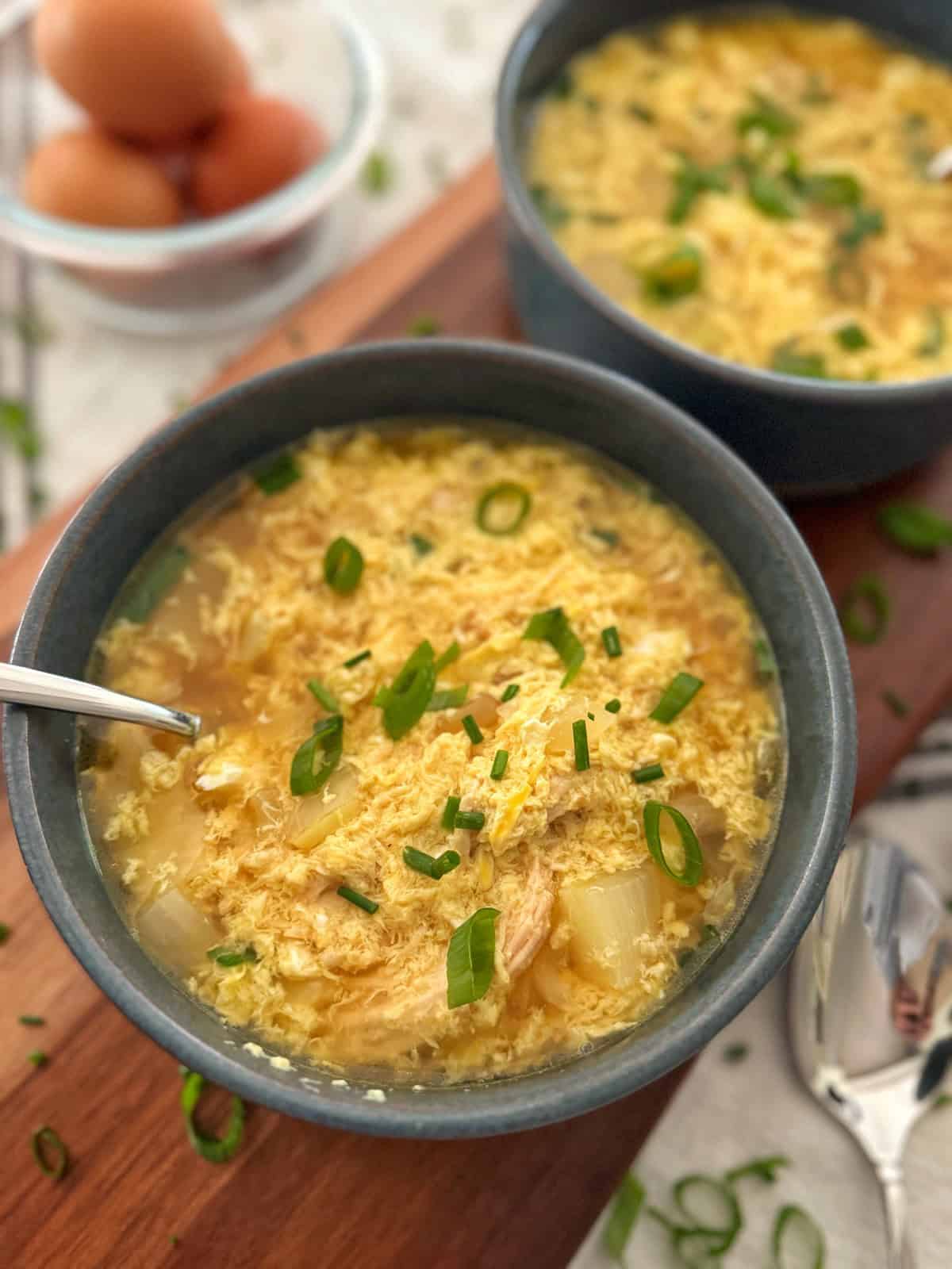 Two bowls of egg drop soup garnished with sliced green onions sit on a wooden surface, with a bowl of eggs and a spoon nearby. The soup contains visible egg ribbons and vegetables.