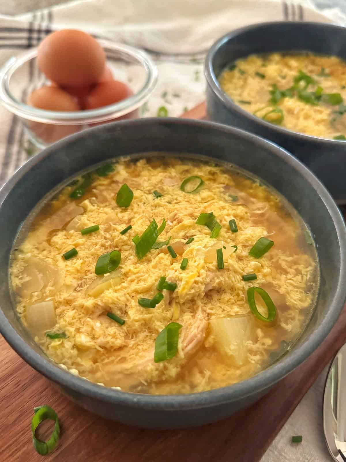 Two bowls of egg drop soup garnished with chopped green onions are placed on a wooden surface. In the background, a glass bowl holds several whole brown eggs.