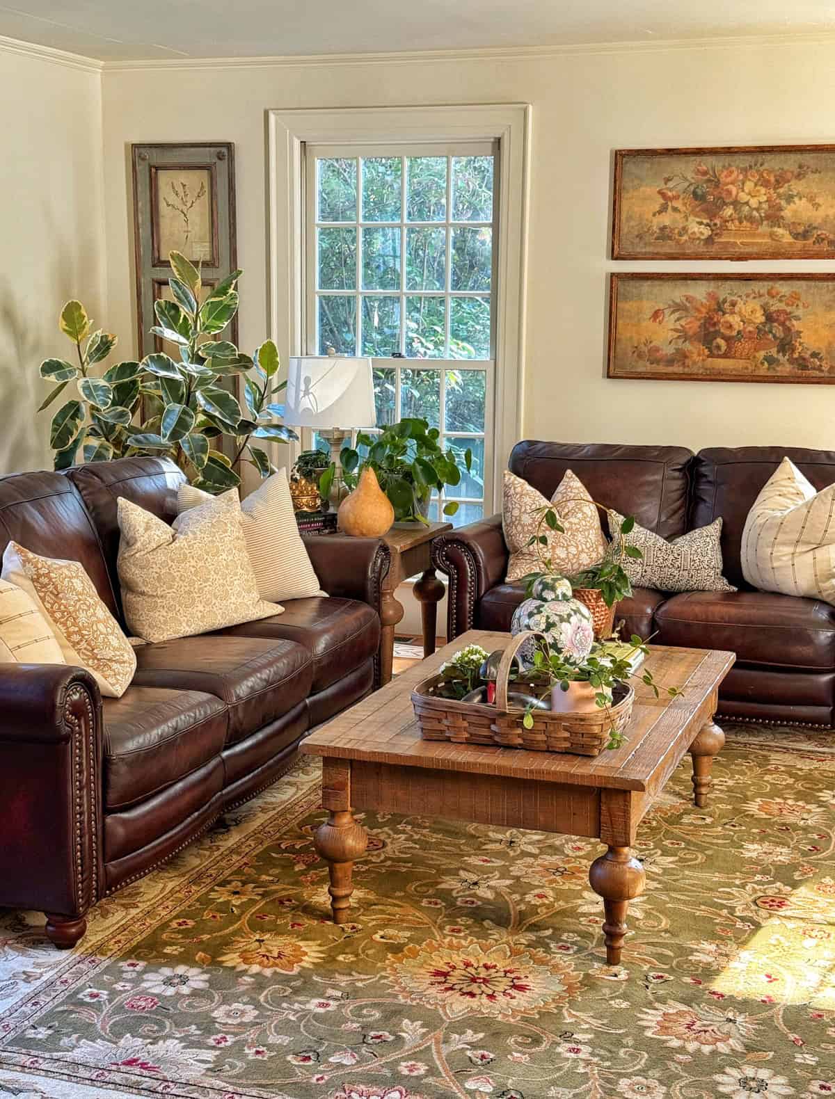 Cozy living room with two dark brown leather sofas, beige and patterned throw pillows, a wooden coffee table with a flower arrangement, potted plants, framed artwork, and a large window letting in natural light.