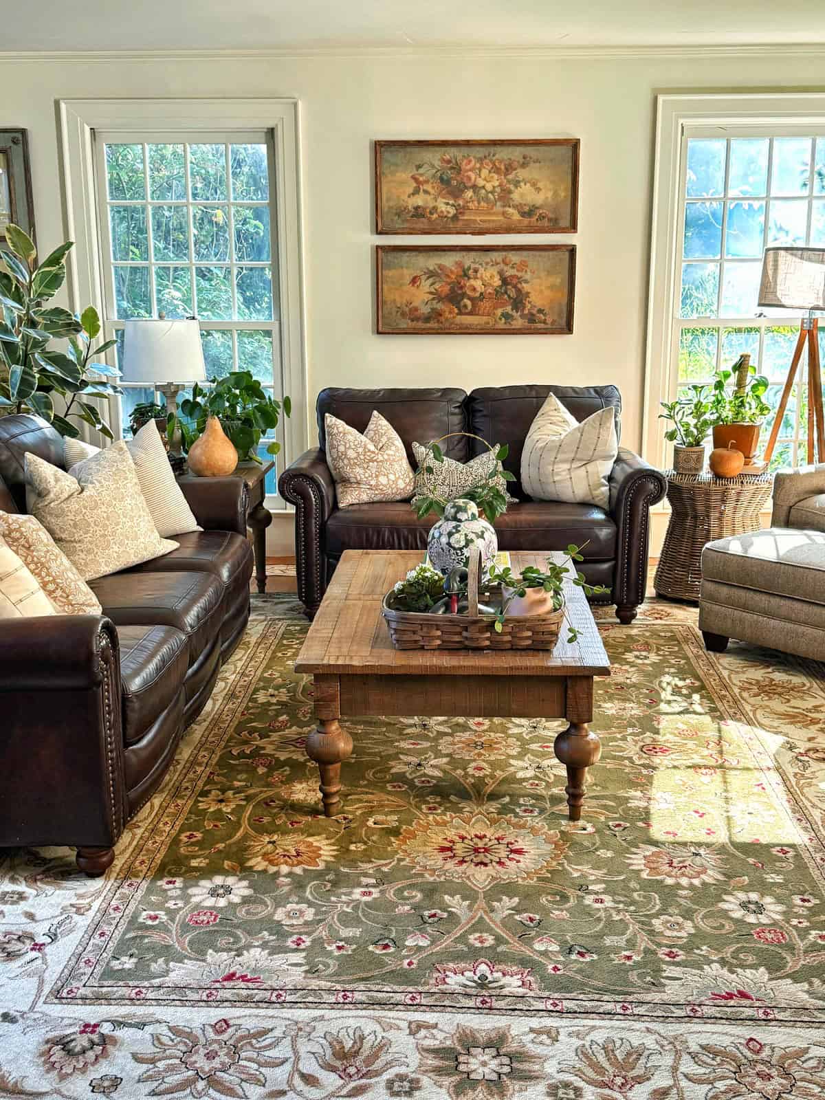 Bright living room with two dark leather sofas, light armchair, wooden coffee table, plants, floral area rug, and framed floral artwork on a cream wall. Large windows let in natural light.