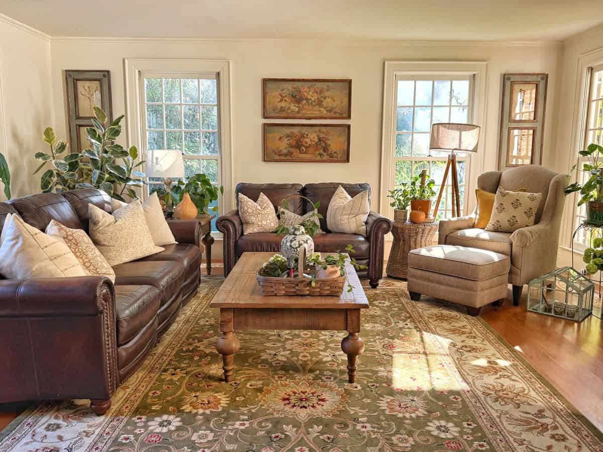 Bright living room with two brown leather sofas, a beige armchair, wooden coffee table, potted plants, and decorative pillows. Large windows let in sunlight, and framed art hangs on the wall above the sofas.