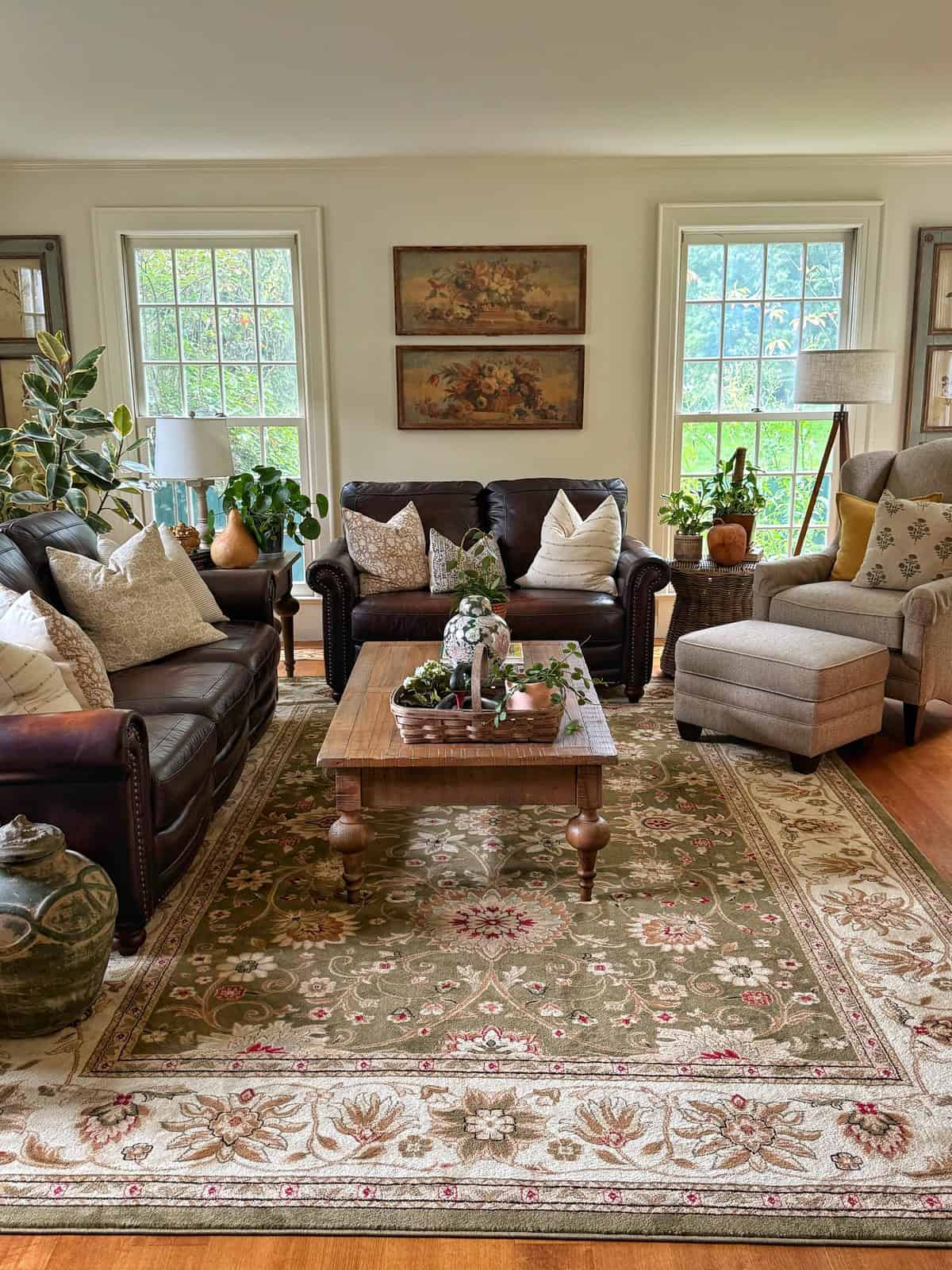 A cozy living room with two sofas, an armchair, and a wooden coffee table on a patterned rug. Plants, decorative pillows, and wall art add warmth, with large windows letting in natural light.