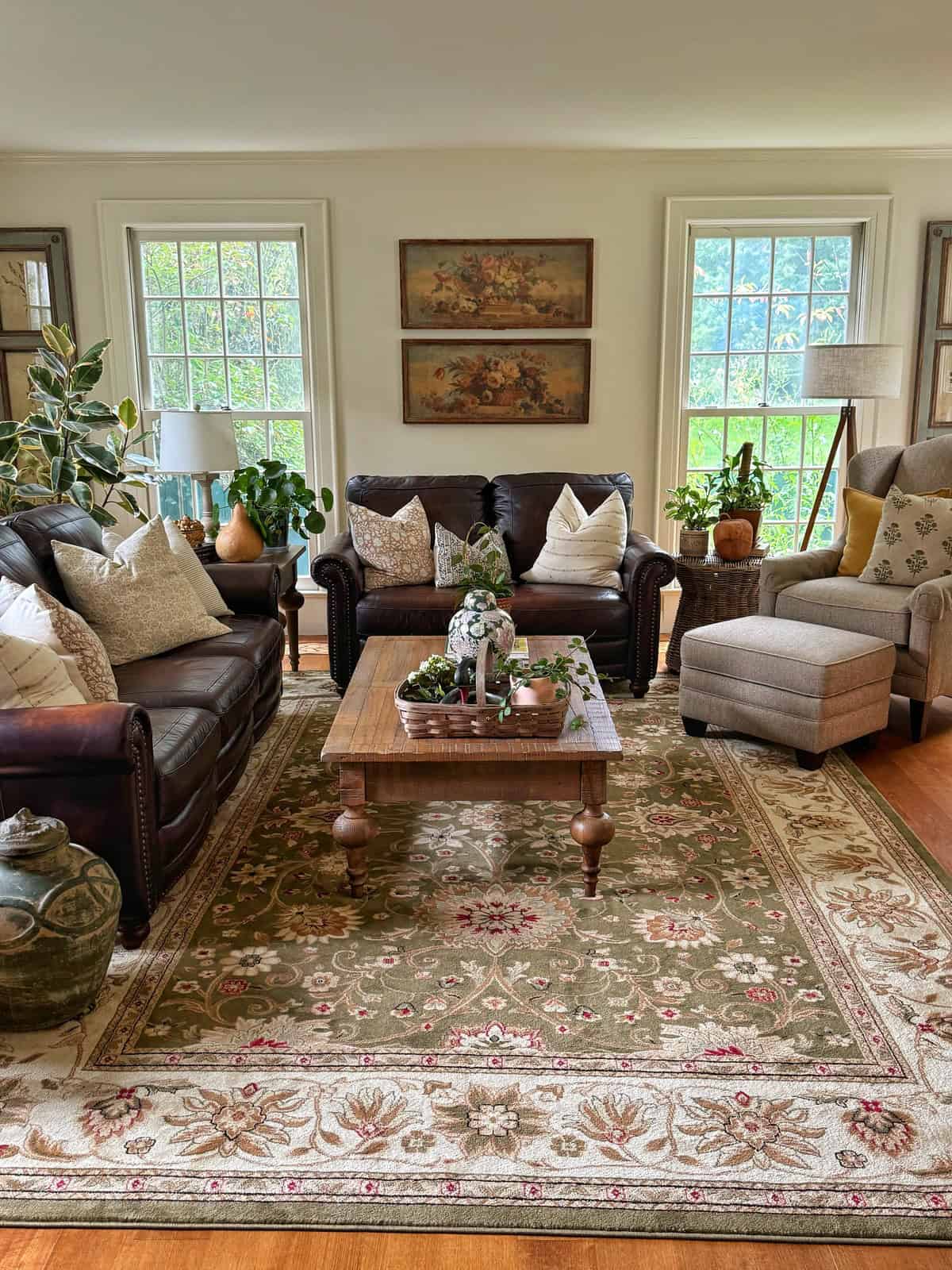 A cozy living room with two sofas, an armchair with an ottoman, a wooden coffee table, decorative pillows, potted plants, and large windows letting in natural light. A patterned rug covers the wooden floor.