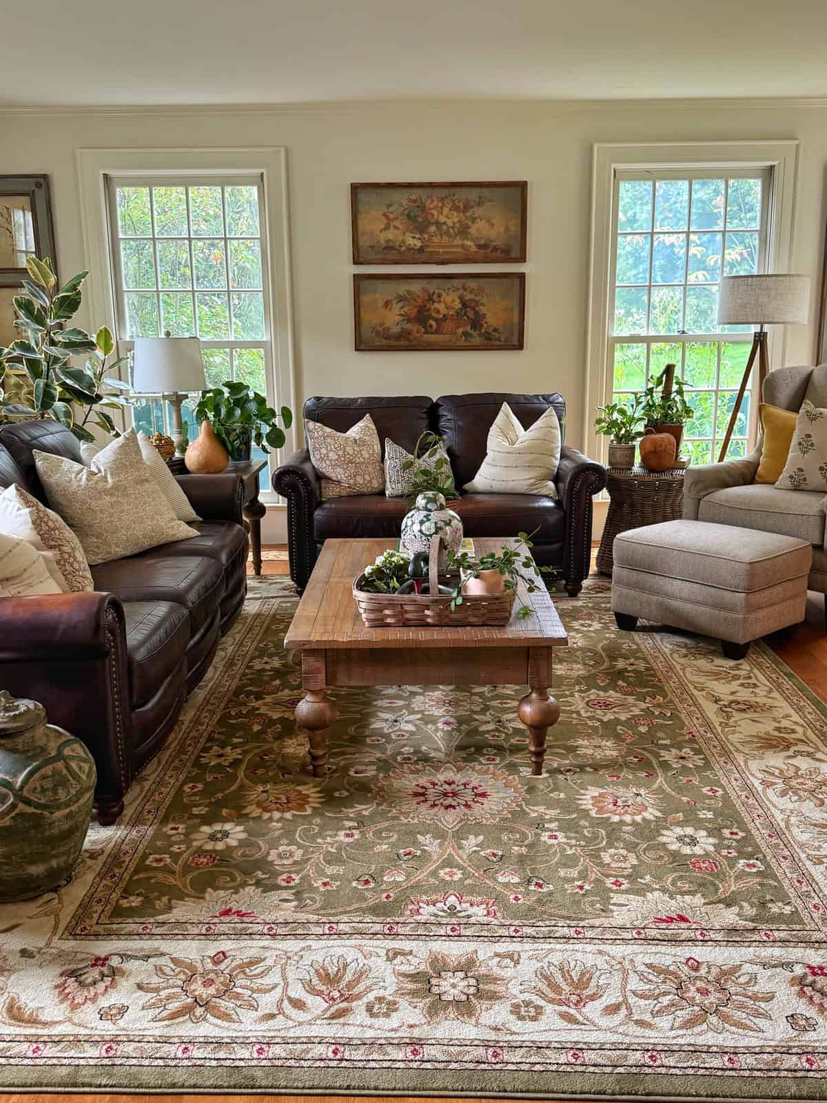 A cozy living room with two dark sofas, a beige armchair, a wooden coffee table, potted plants, and floral artwork on the wall. Large windows let in natural light, and a patterned rug covers the wooden floor.