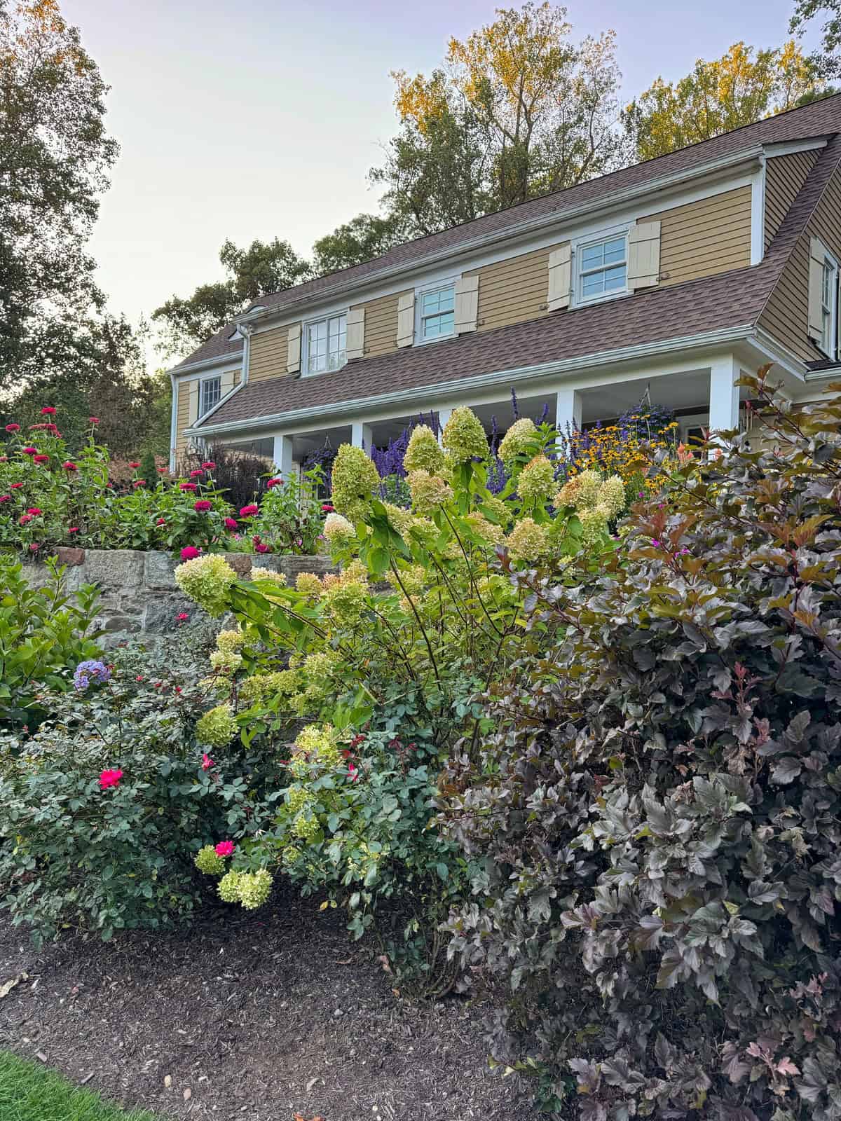 A large beige house with white trim and shutters stands behind a lush garden filled with blooming flowers and shrubs, bordered by a stone wall and surrounded by tall trees under a clear sky.