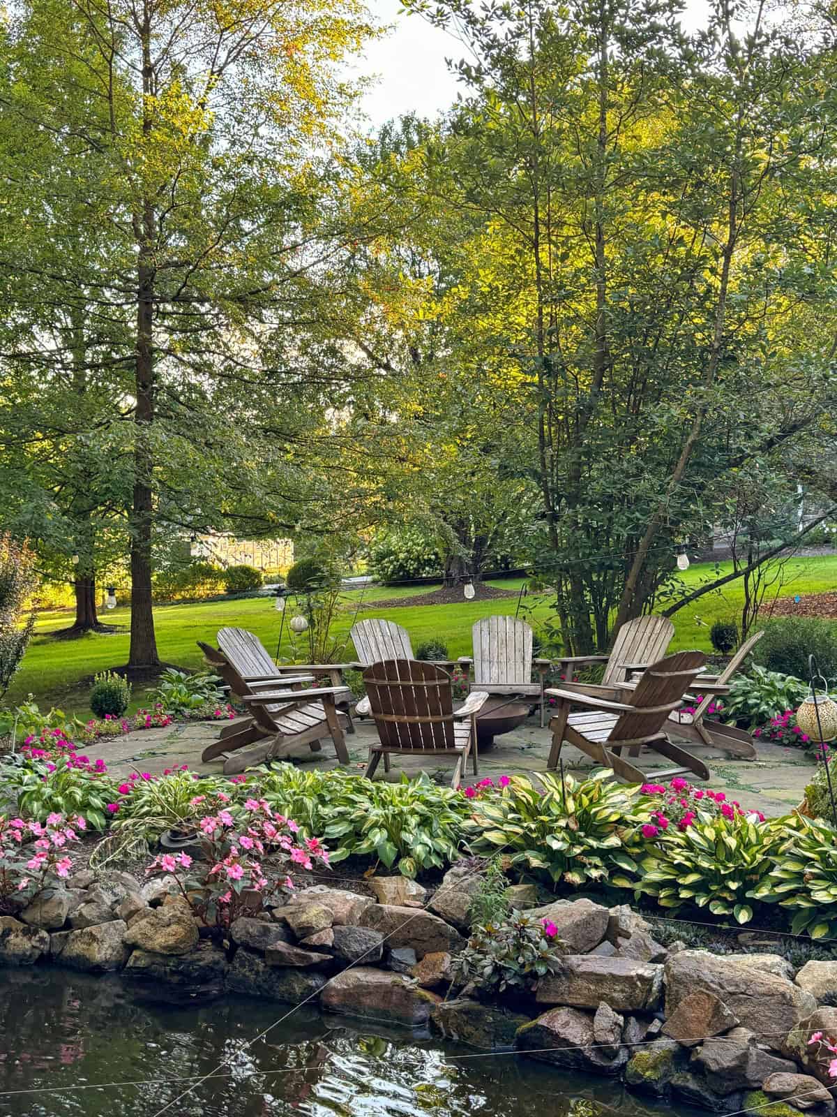 A group of wooden Adirondack chairs surrounds a fire pit on a stone patio, bordered by blooming pink flowers and rocks, with lush green trees and grass in the background.