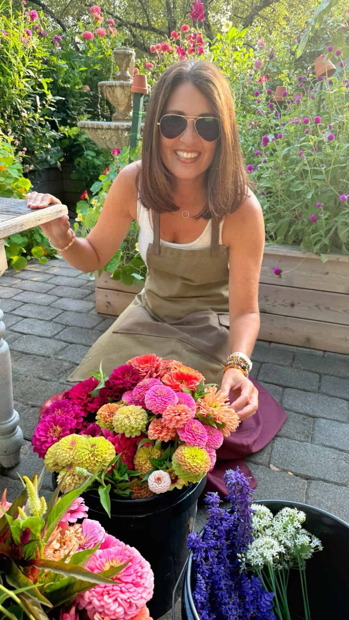 A woman wearing sunglasses and an apron kneels on a patio, smiling while arranging colorful flowers in a black bucket. She is surrounded by lush greenery and blooming garden beds.