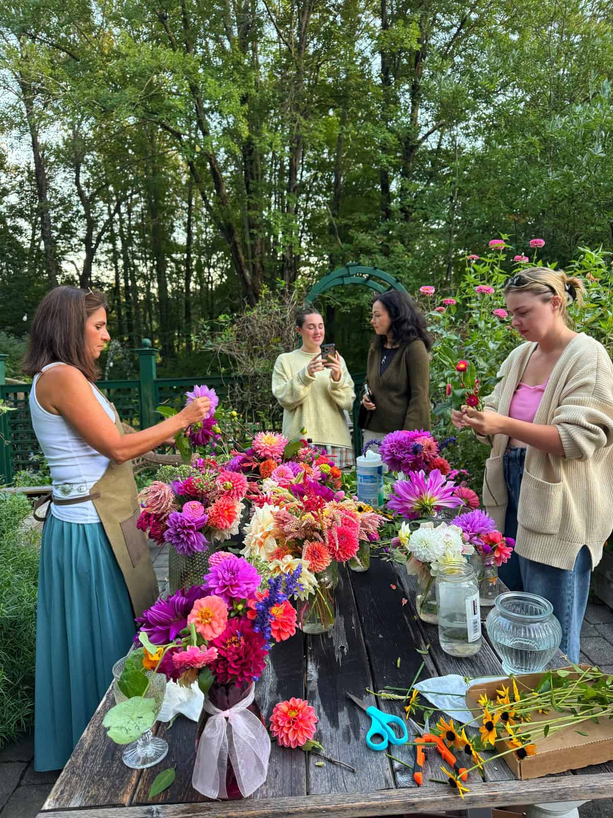Four women (including stacy ling) stand around a rustic wooden table outdoors, arranging colorful flowers in jars. The table is filled with vibrant dahlias, zinnias, and sunflowers. Trees and greenery surround the scene, creating a peaceful cut flower garden setting.