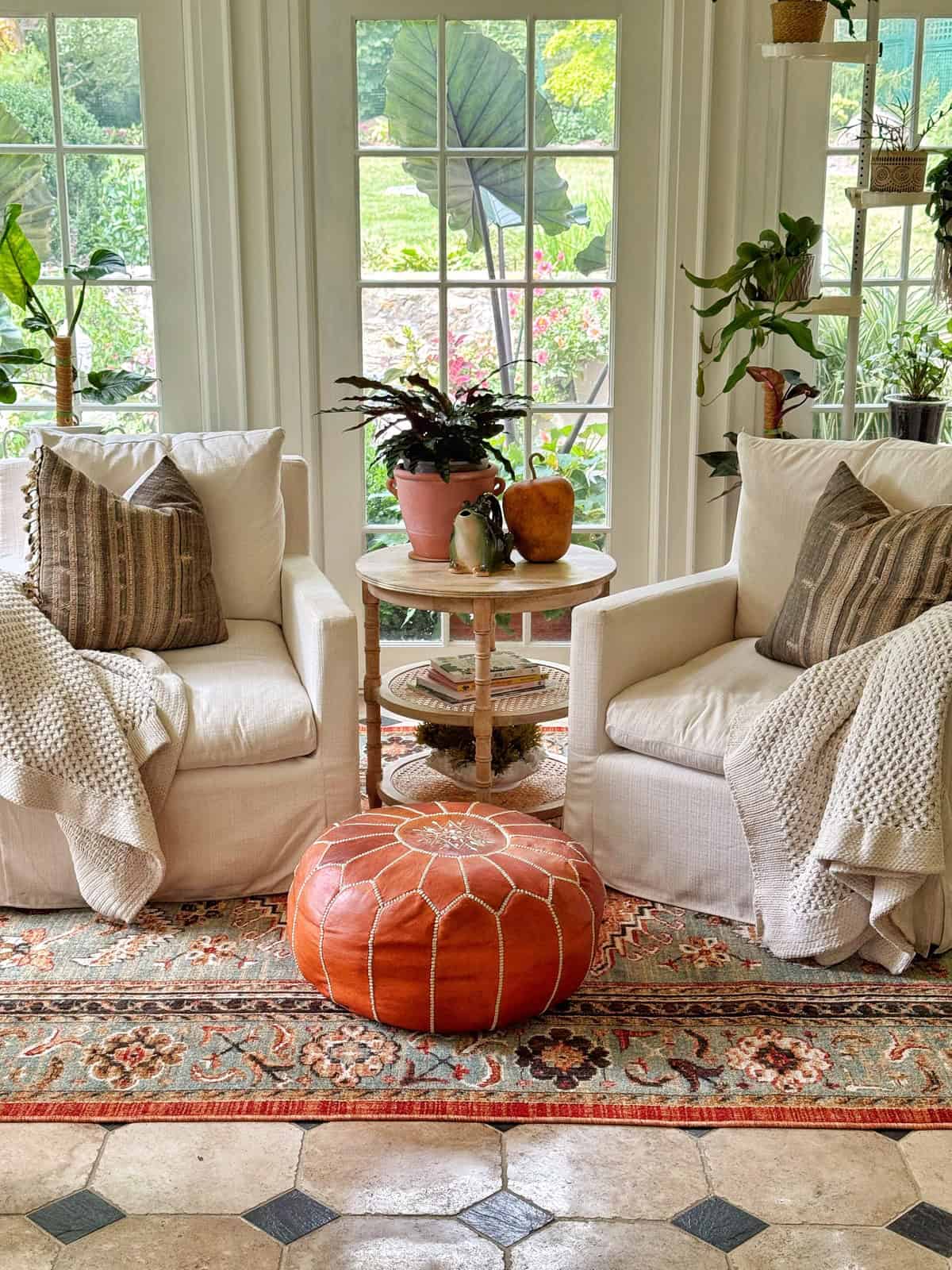 Two cozy white armchairs with striped pillows and blankets sit by a small round table with plants and decorative items. A tan leather pouf rests on a patterned rug in front of large windows overlooking a garden.