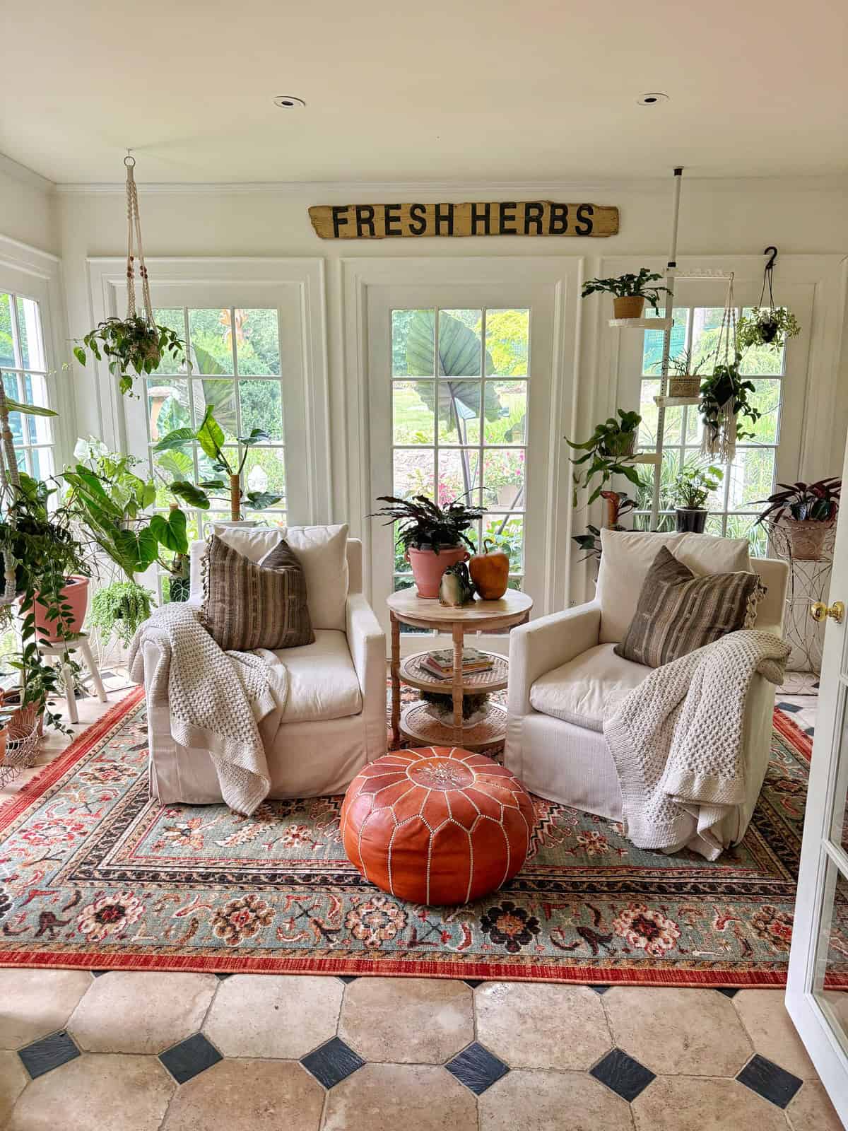 A cozy sunroom with two white armchairs, a round leather ottoman, potted houseplants, hanging greenery, a patterned rug, and a sign reading “FRESH HERBS” above the windows. Sunlight streams in through large windows.