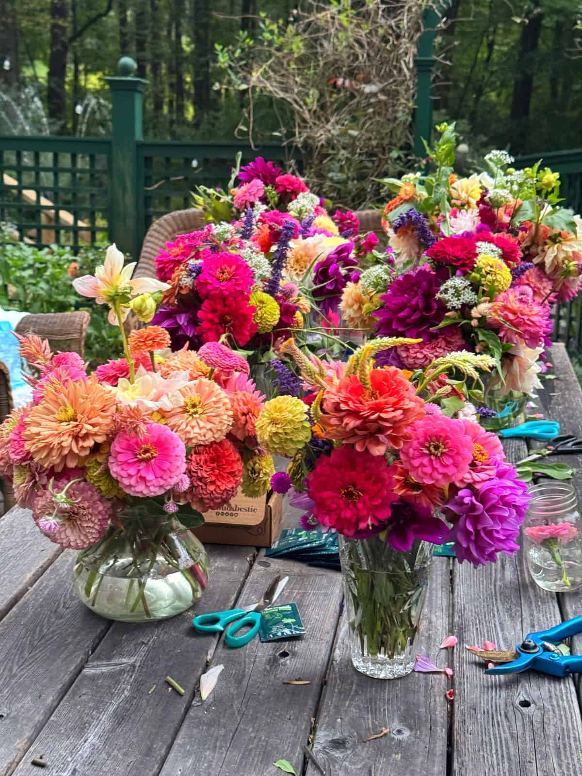 Colorful bouquets of assorted flowers in glass vases sit on a rustic wooden table outdoors, surrounded by scissors, flower stems, and leaves, with a green fence and trees in the background.