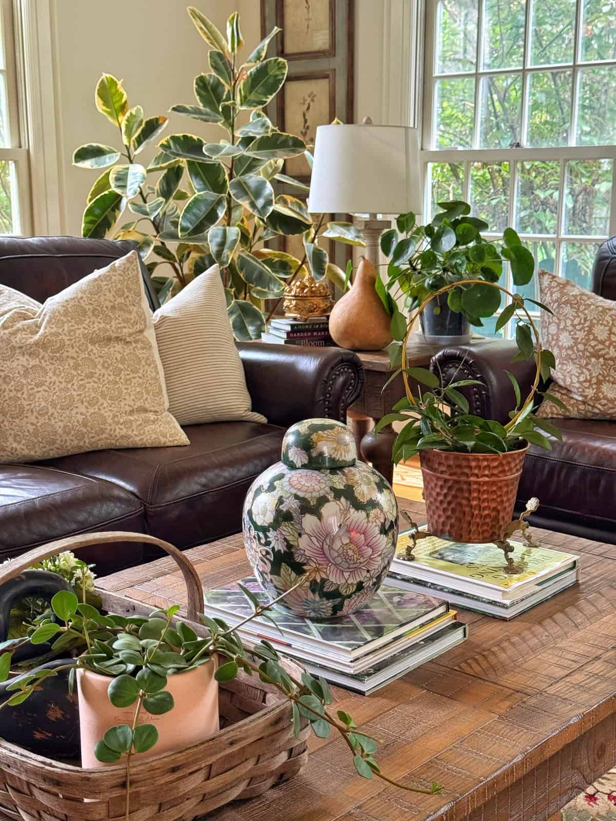 A cozy living room with brown leather sofas, patterned pillows, leafy potted plants, a floral ceramic jar, and stacked books on a wooden coffee table, with large windows letting in natural light.