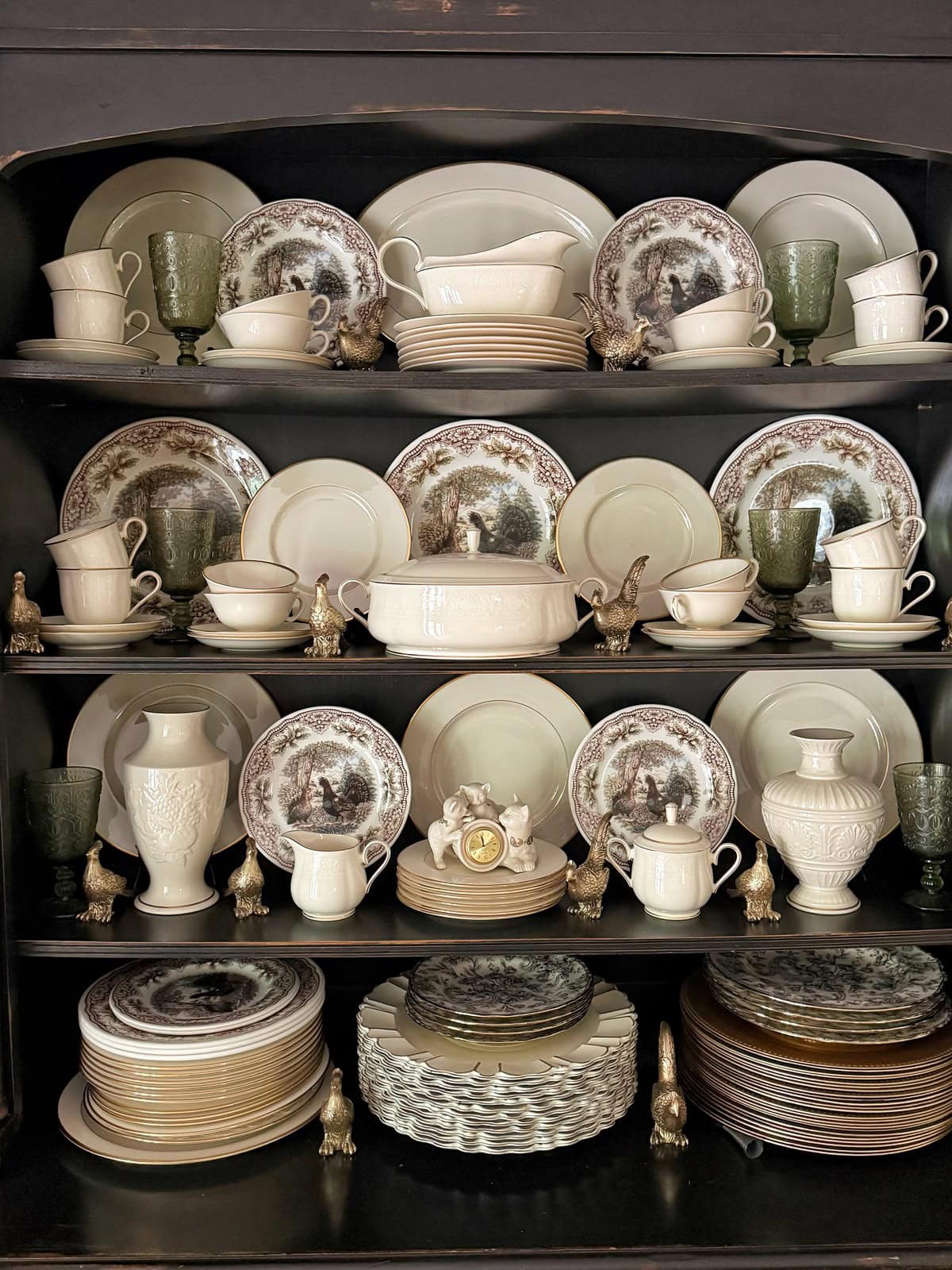 A black hutch filled with neatly arranged vintage dishes, cups, goblets, and serving pieces. Plates with pastoral scenes, cream-colored ceramics, and small gold bird figurines are displayed on three shelves.