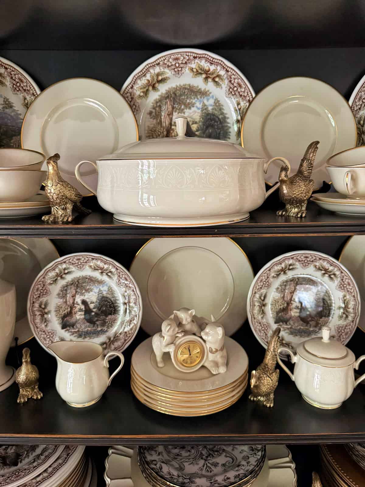 A china cabinet shelf displaying vintage china plates, cups, and a tureen, accented with gold trim and nature scenes. Brass bird figurines and a small ceramic pig clock decorate the arrangement.