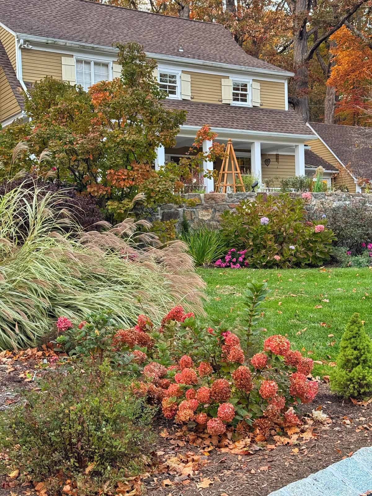 A two-story house with tan siding and white trim is surrounded by a well-maintained garden with blooming flowers, green grass, shrubs, and autumn leaves scattered on the ground.