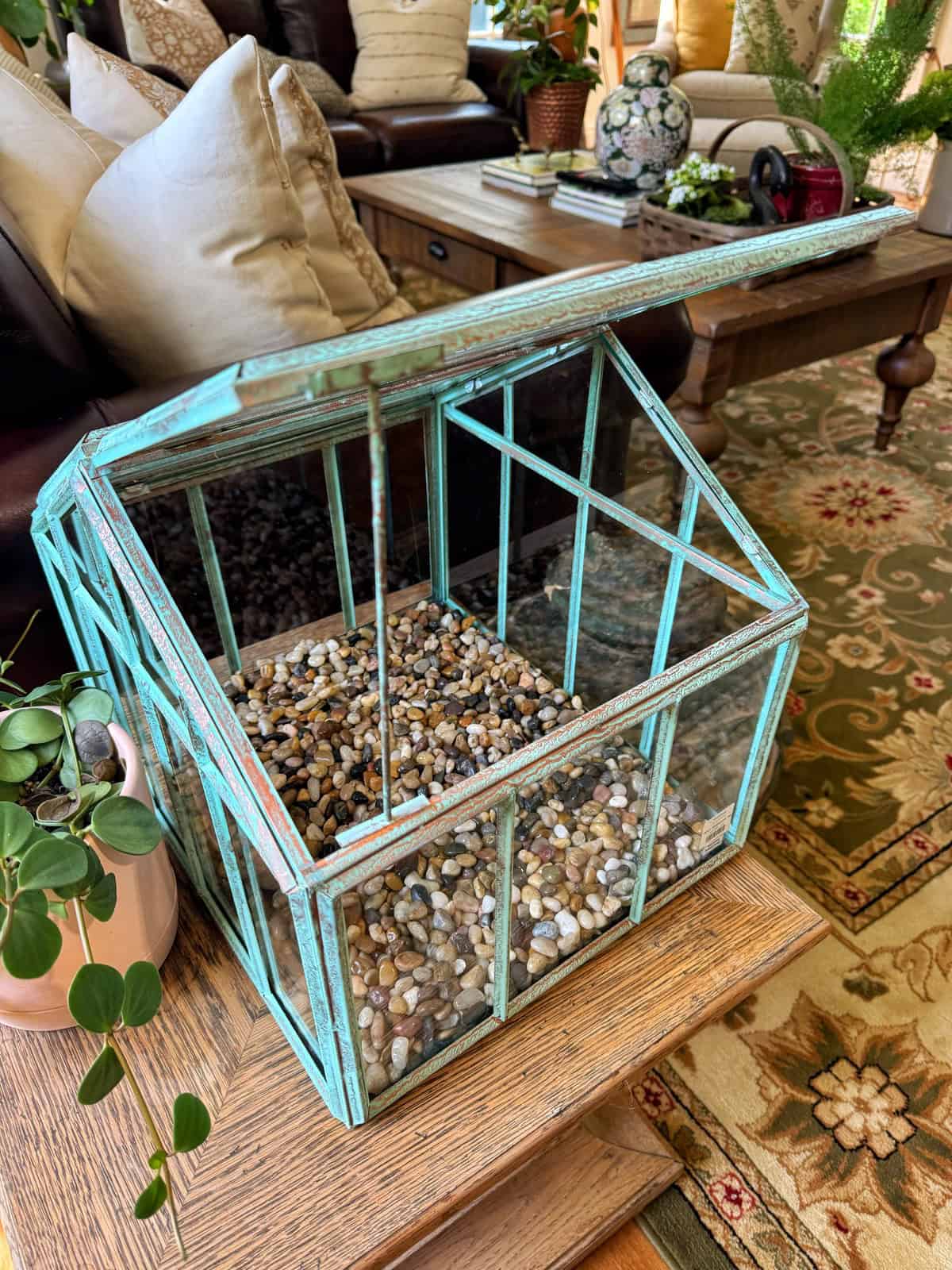 A glass and turquoise metal terrarium with its lid open sits on a table, partially filled with small pebbles. Nearby is a potted plant, and a cozy living room setting is visible in the background.
