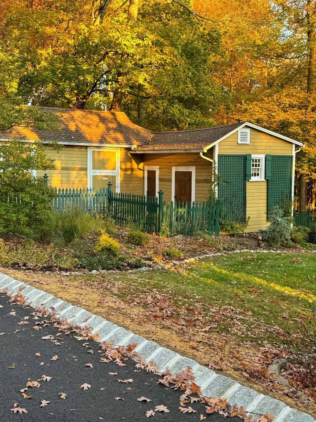 A small yellow house with green shutters and a green picket fence sits amid autumn trees, fallen leaves covering the ground and sunlight casting a warm glow over the scene.
