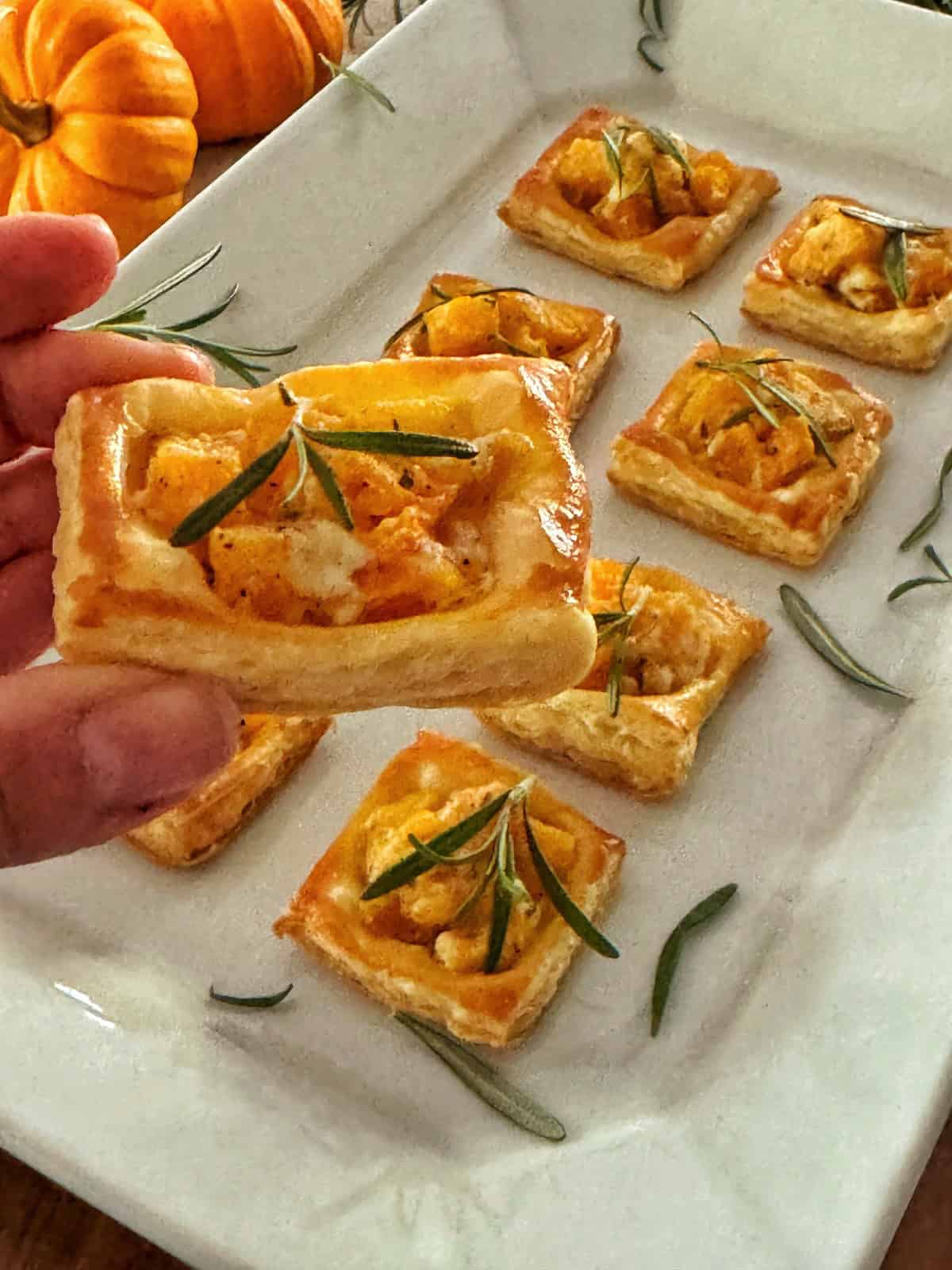 A hand holds a square pumpkin puff pastry topped with diced roasted squash and a sprig of rosemary. Several similar pastries are arranged on a white plate, with rosemary leaves scattered around. Small pumpkins are in the background.