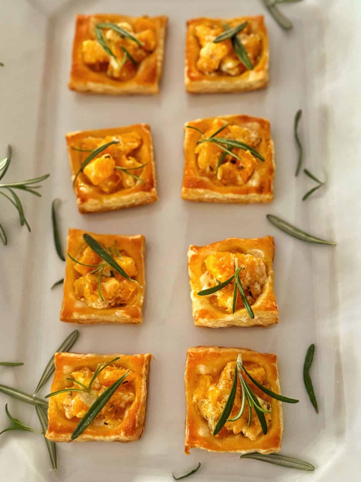 Eight square puff pastry appetizers topped with yellow roasted vegetables and garnished with green rosemary sprigs are arranged neatly on a white tray, with extra rosemary scattered around.