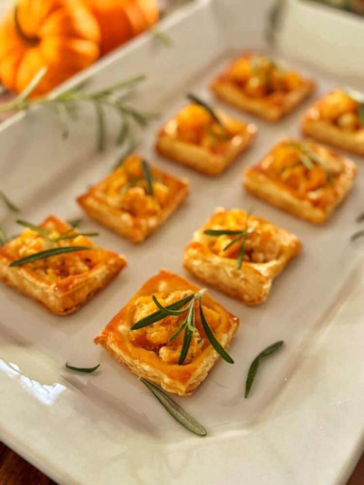 Eight square pumpkin puff pastry bites topped with orange filling and sprigs of rosemary are arranged neatly on a white platter. A small pumpkin and more rosemary are visible in the blurred background.