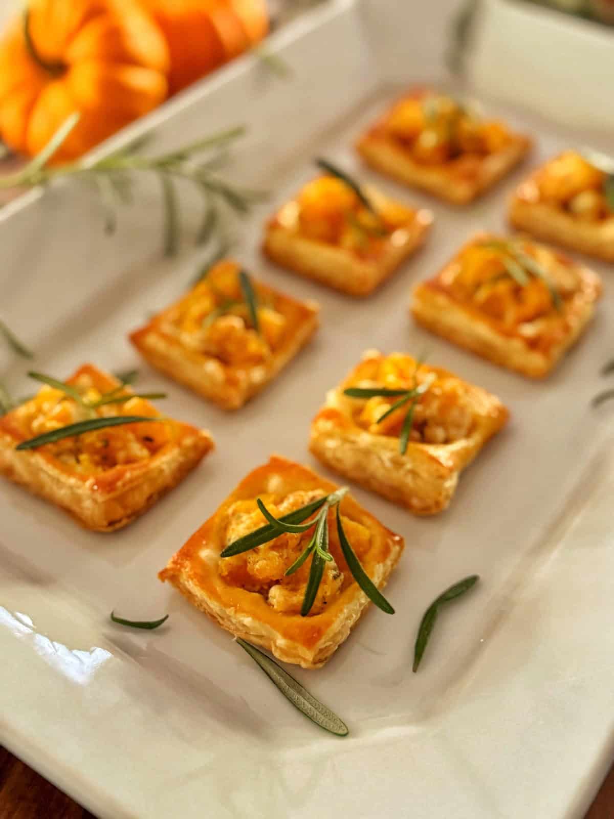 Eight square pumpkin puff pastry bites topped with orange filling and sprigs of rosemary are arranged neatly on a white platter. A small pumpkin and more rosemary are visible in the blurred background.