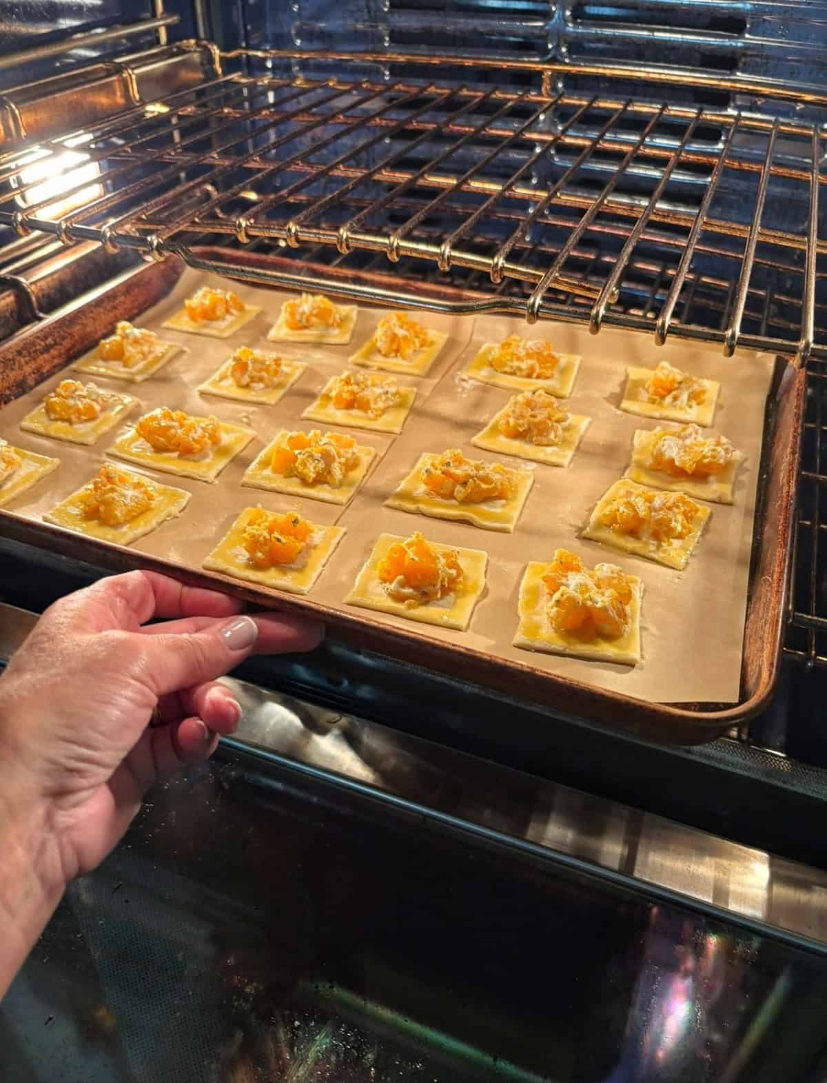 A hand places a baking sheet lined with parchment paper and rows of small pastry squares topped with an orange filling into an oven.