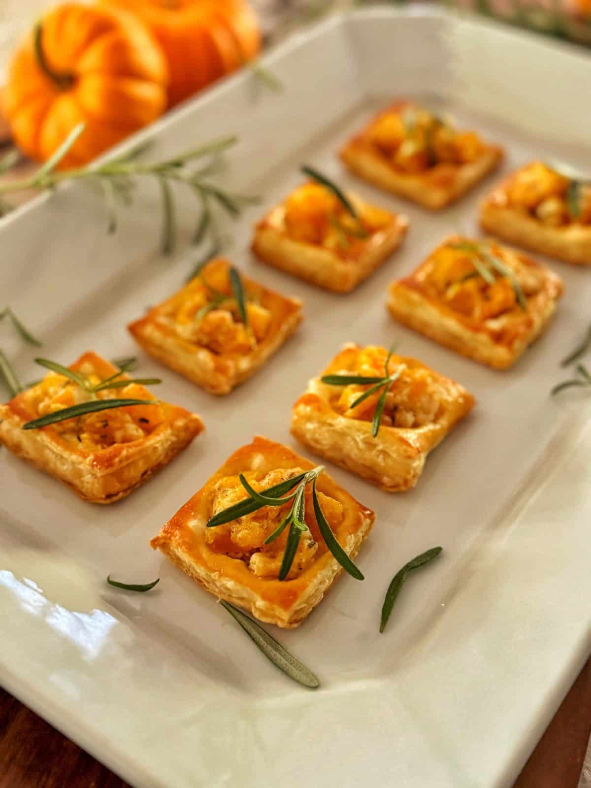 A white platter with small square pumpkin puff pastry appetizers topped with orange squash or pumpkin and fresh rosemary, with mini pumpkins and sprigs of rosemary in the background.