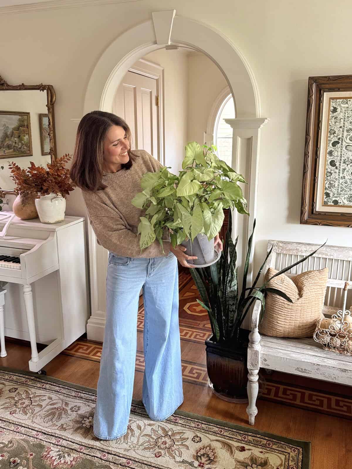 A woman in a beige sweater and wide-leg jeans stands indoors, smiling and holding a potted leafy plant. She is in a cozy, decorated room with a piano, plants, a rug, and framed artwork on the wall.