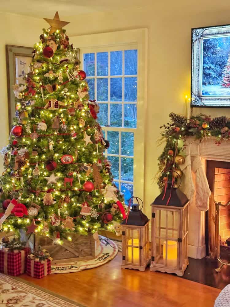 A decorated Christmas tree with lights and ornaments stands by a window. Nearby, a fireplace is adorned with stockings and garland, and two lanterns sit on the floor, creating a warm, festive atmosphere.