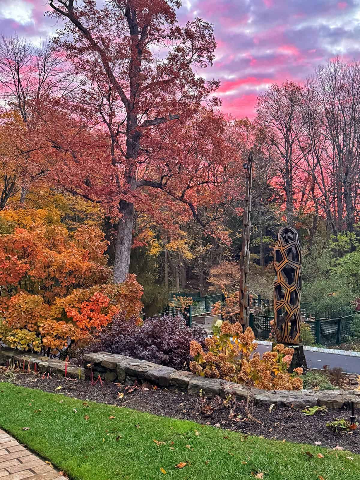 A vibrant autumn garden with colorful trees, a stone-bordered flower bed, and a tall, honeycomb-shaped wooden sculpture. The sky glows pink and purple at sunset, casting warm light over the scene.