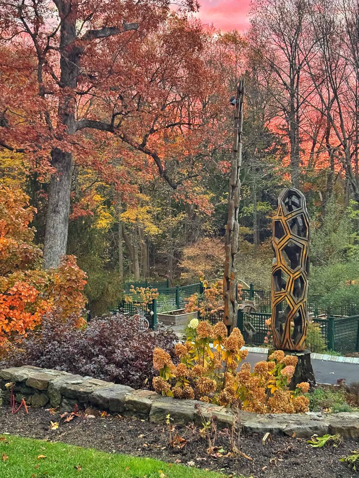 A garden with autumn trees, colorful leaves, stone edging, and tall abstract sculptures stands under a vivid pink and orange sunset sky. A green fence is visible in the background.